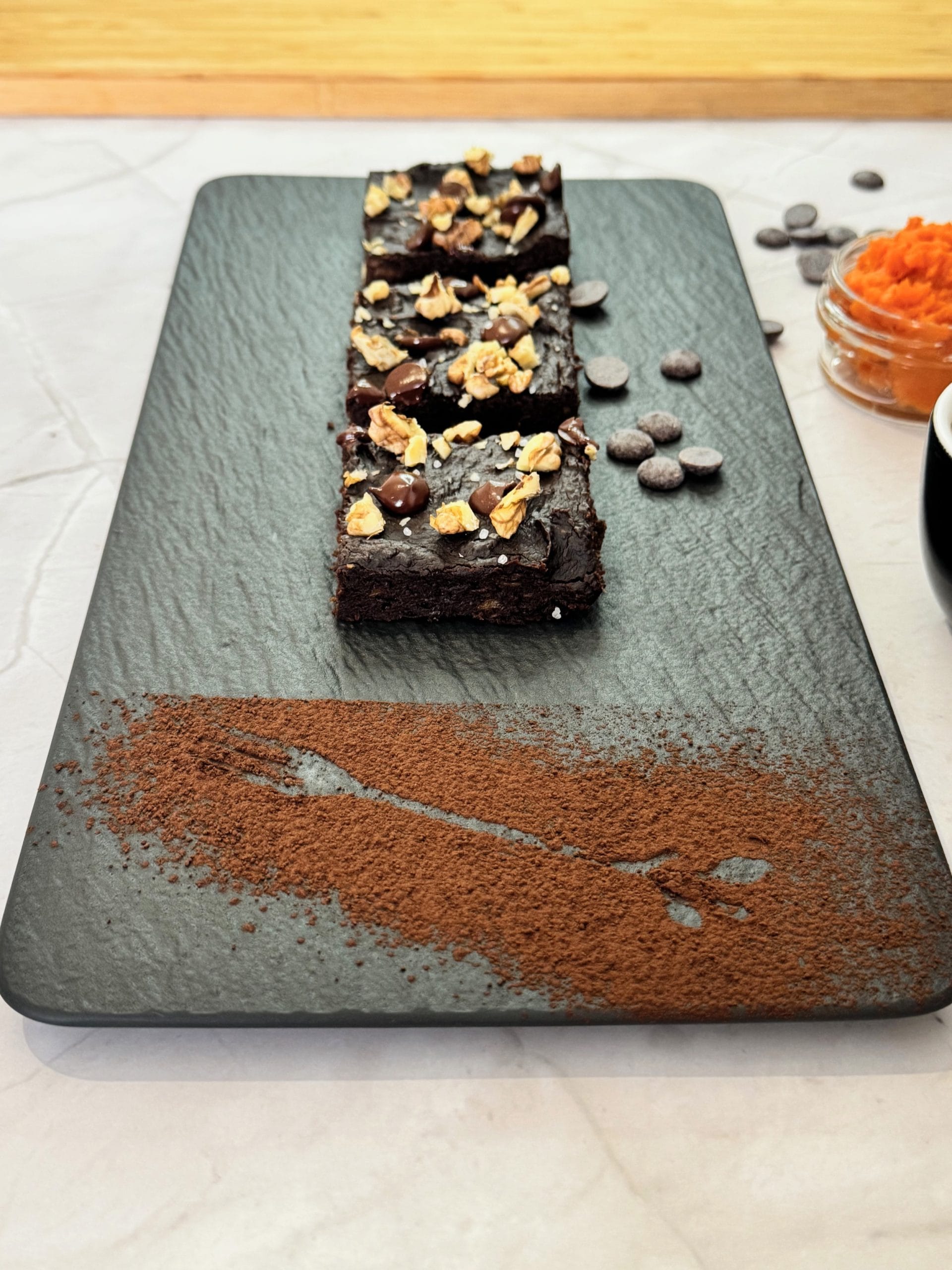 Felii de brownie cu cartof dulce, cu nuci, ciocolată și fulgi de sare, aranjate pe un platou de ardezie presărat cu cacao, alături de cartof dulce piure și cafea