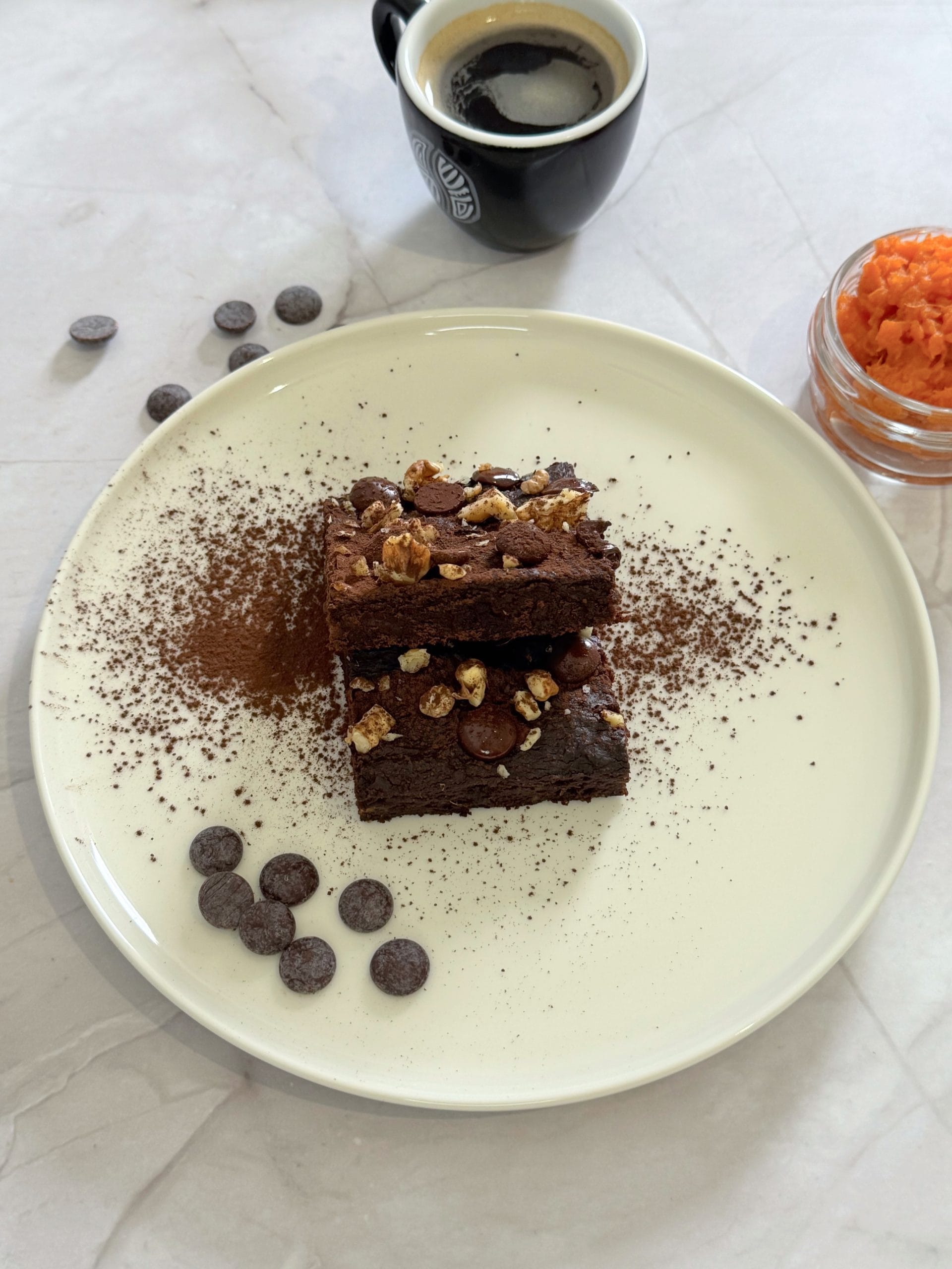 Două bucăți de brownie cu cartof dulce, stivuite pe o farfurie, presărate cu cacao, nuci și fulgi de ciocolată, alături de o cafea și un bol cu piure de cartof dulce
