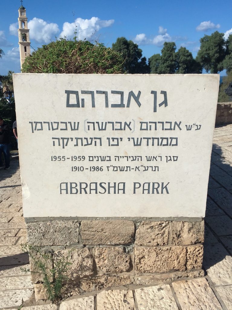 Placă din piatră cu numele parcului Abrasha &icirc;n ebraică și engleză, cu vegetație și un turn de biserică &icirc;n fundal