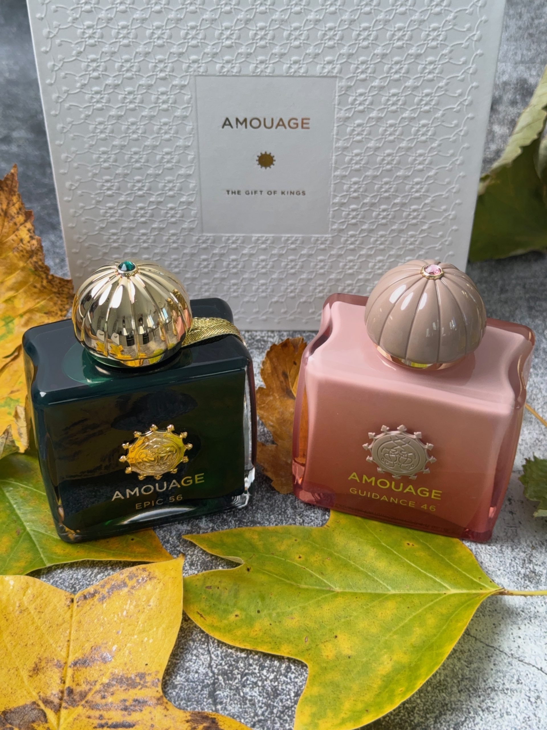 Sticlele parfumurilor Amouage Epic 56, verde închis, și Guidance 46, roz, alături de ambalajul alb Amouage și frunze colorate de toamnă