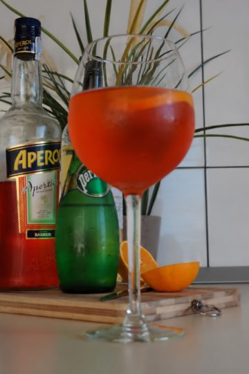 Glass of Aperol Spritz with Aperol and sparkling water bottles behind it and orange pieces on a wooden board