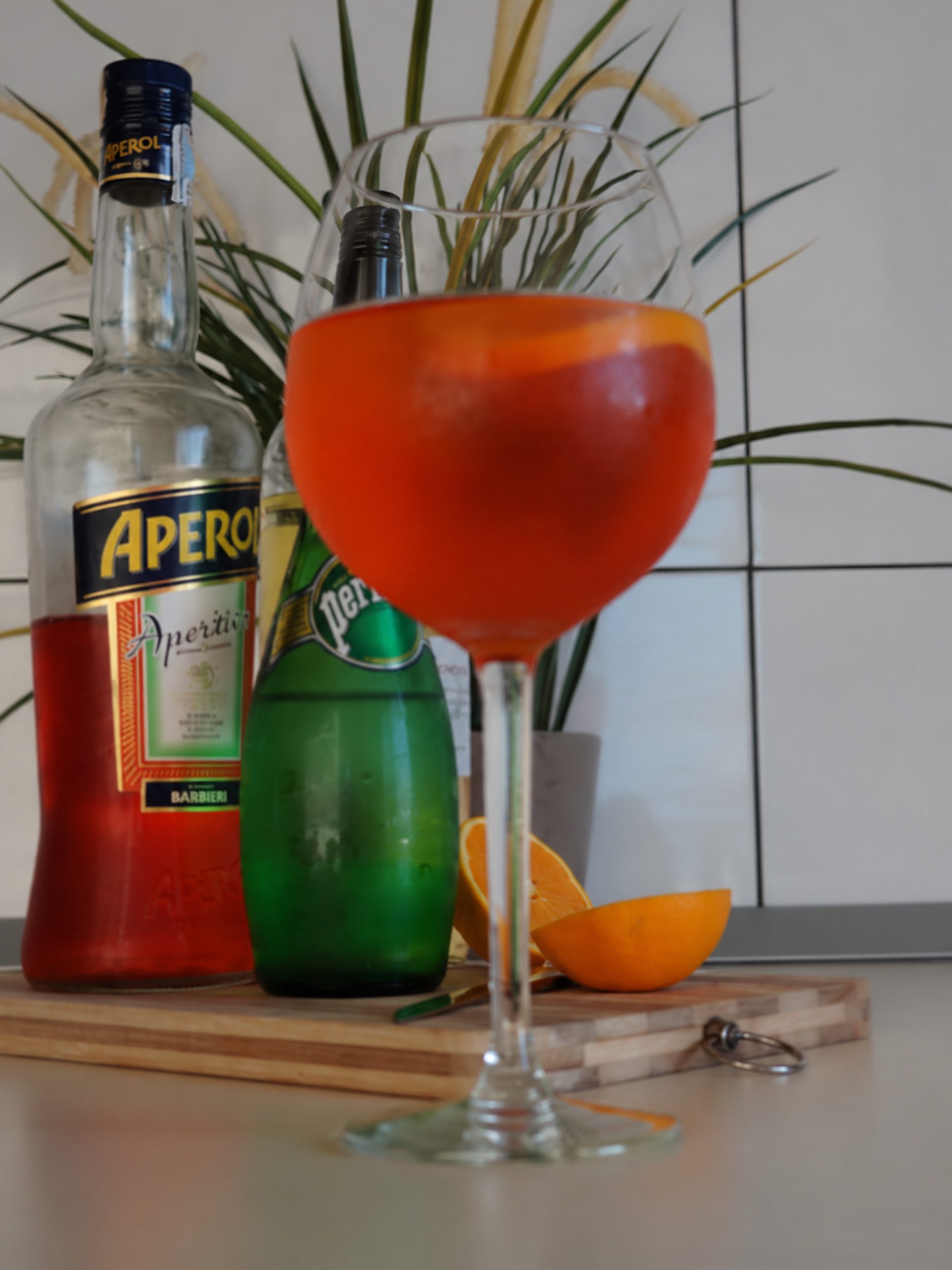 Glass of Aperol Spritz with Aperol and sparkling water bottles behind it and orange pieces on a wooden board