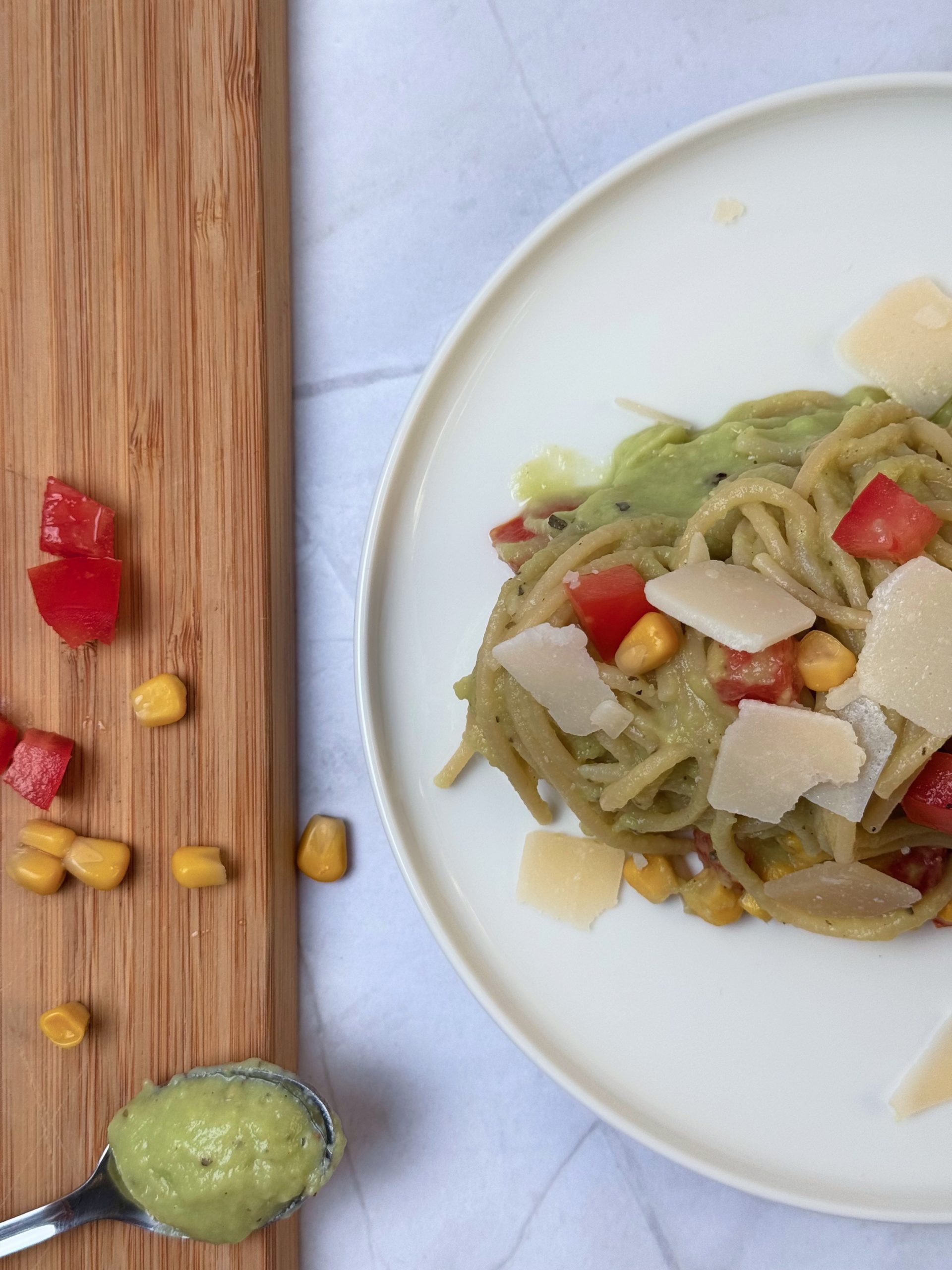 Spaghete cu sos de avocado, porumb, roșii și parmezan, alături de o lingură cu sos cremos și un tocător cu legume