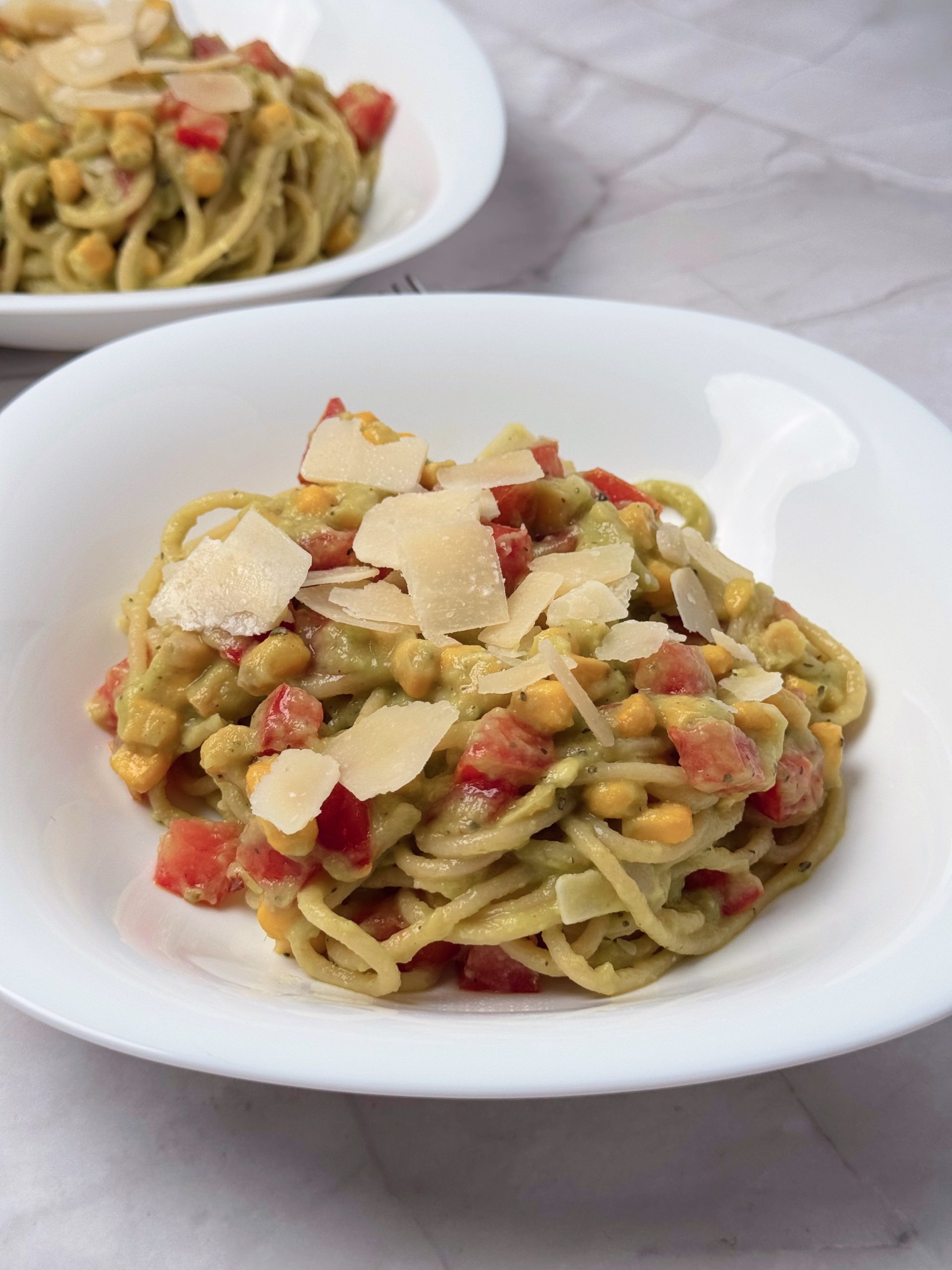 Prim-plan cu spaghete în sos cremos de avocado, cu boabe de porumb, roșii tăiate cubulețe și fulgi de parmezan