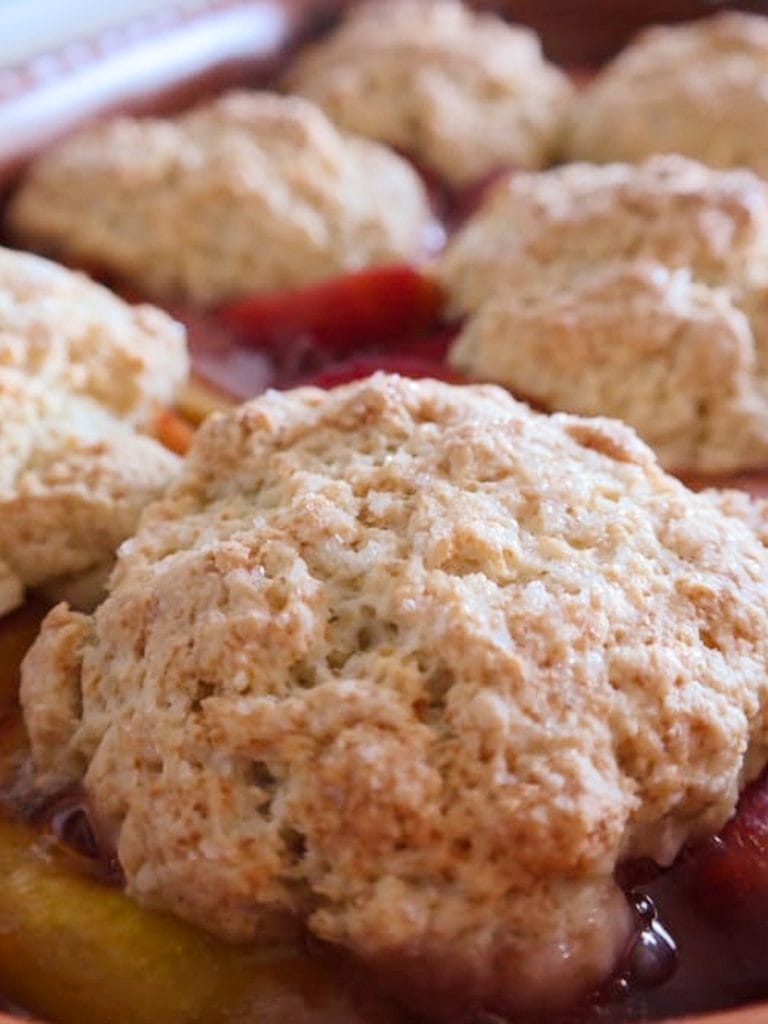 Baked peach cobbler in a terracotta dish with crisp golden biscuit topping over bubbling peaches