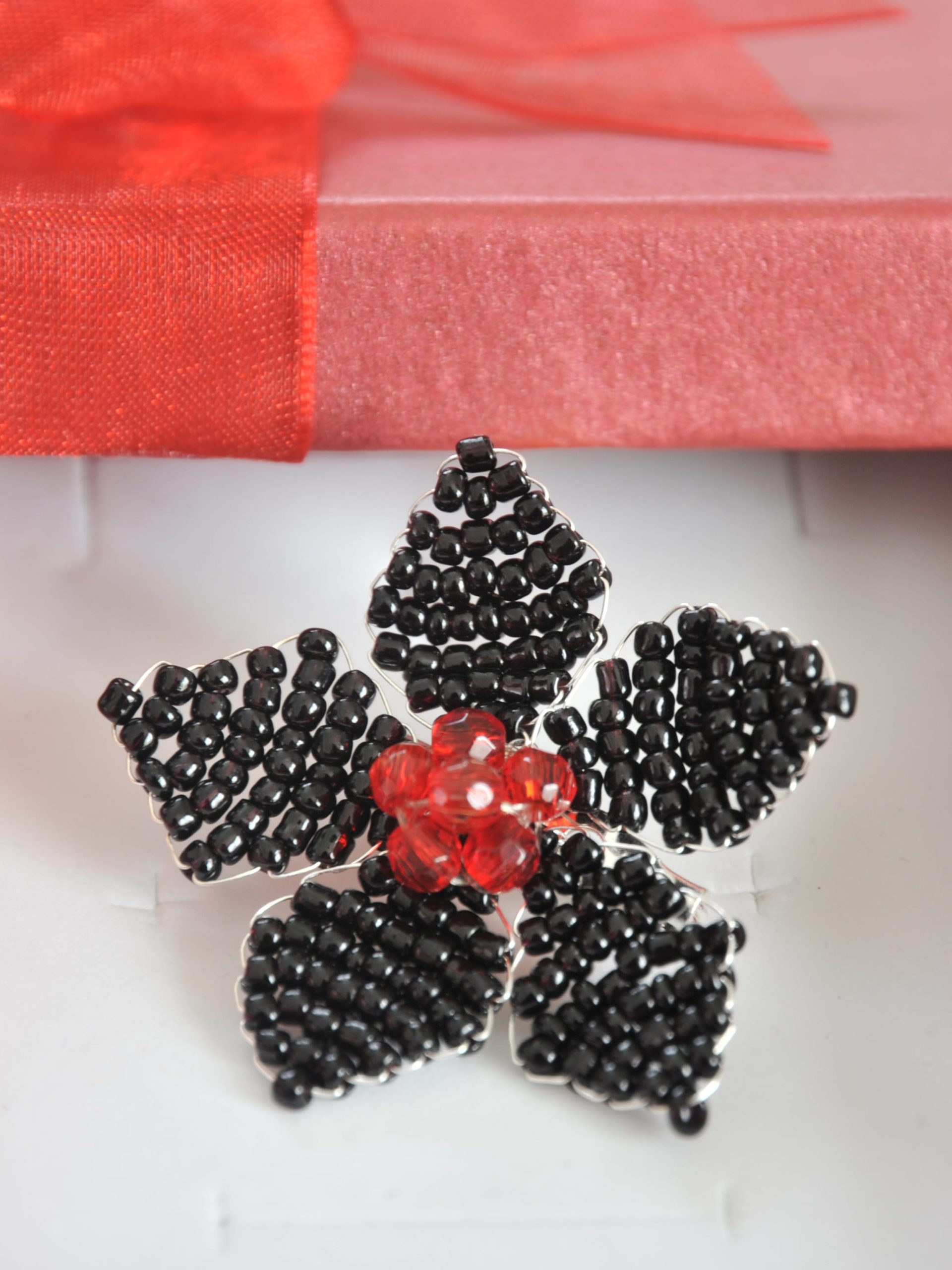 Handmade flower-shaped brooch with five petals of black beads set in silver wire and a center of red crystal beads, in a red gift box