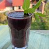 Pahar cu smoothie violet, decorat cu frunze de mentă, pe un prosop verde, cu case și vegetație în fundal