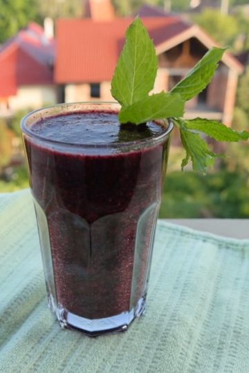 Pahar cu smoothie violet, decorat cu frunze de mentă, pe un prosop verde, cu case și vegetație în fundal