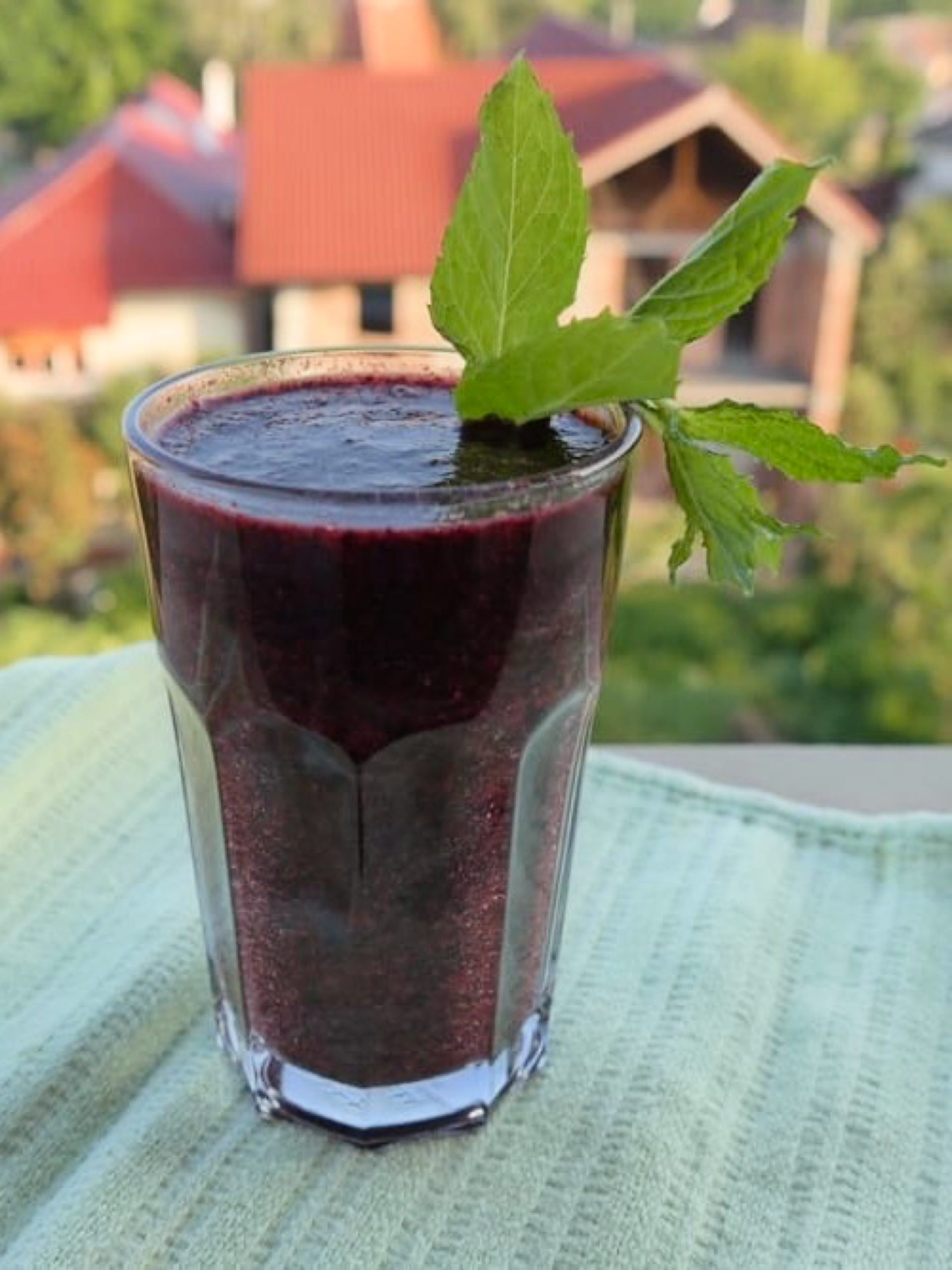 Pahar cu smoothie violet, decorat cu frunze de mentă, pe un prosop verde, cu case și vegetație &icirc;n fundal