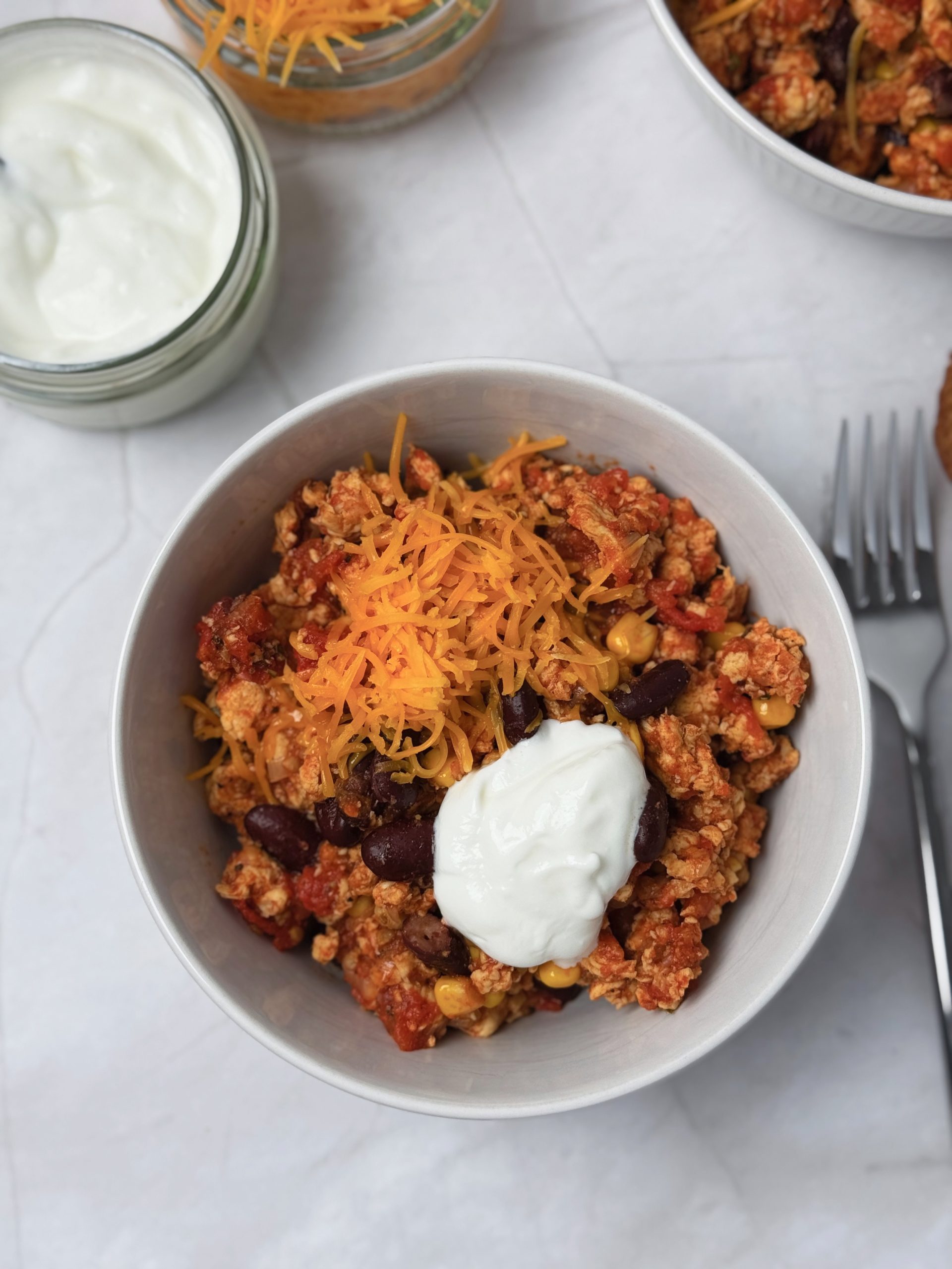 Bowl of chicken chilli con carne with kidney beans and sweetcorn, topped with grated cheddar cheese and sour cream