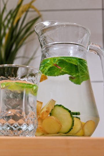 Glass pitcher and tumbler of infused water with mint leaves and slices of cucumber, lemon and ginger