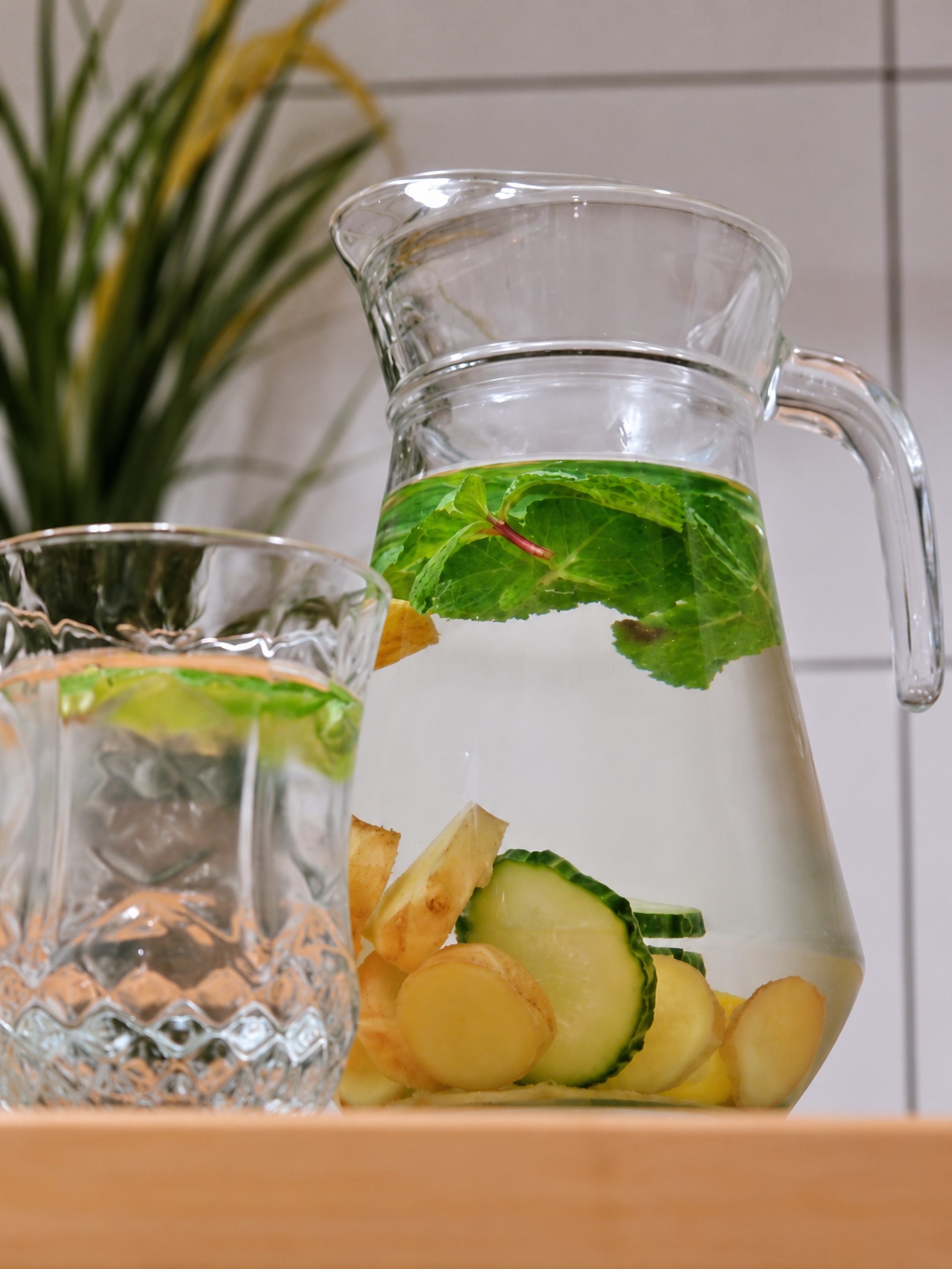 Glass pitcher and tumbler of infused water with mint leaves and slices of cucumber, lemon and ginger