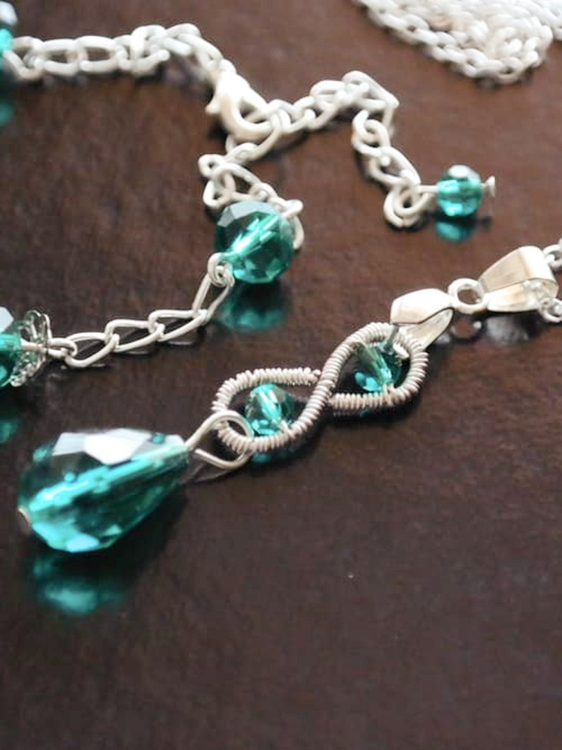 Wire-wrapped infinity pendant with an emerald green teardrop crystal next to a silver chain bracelet with green beads on a dark surface