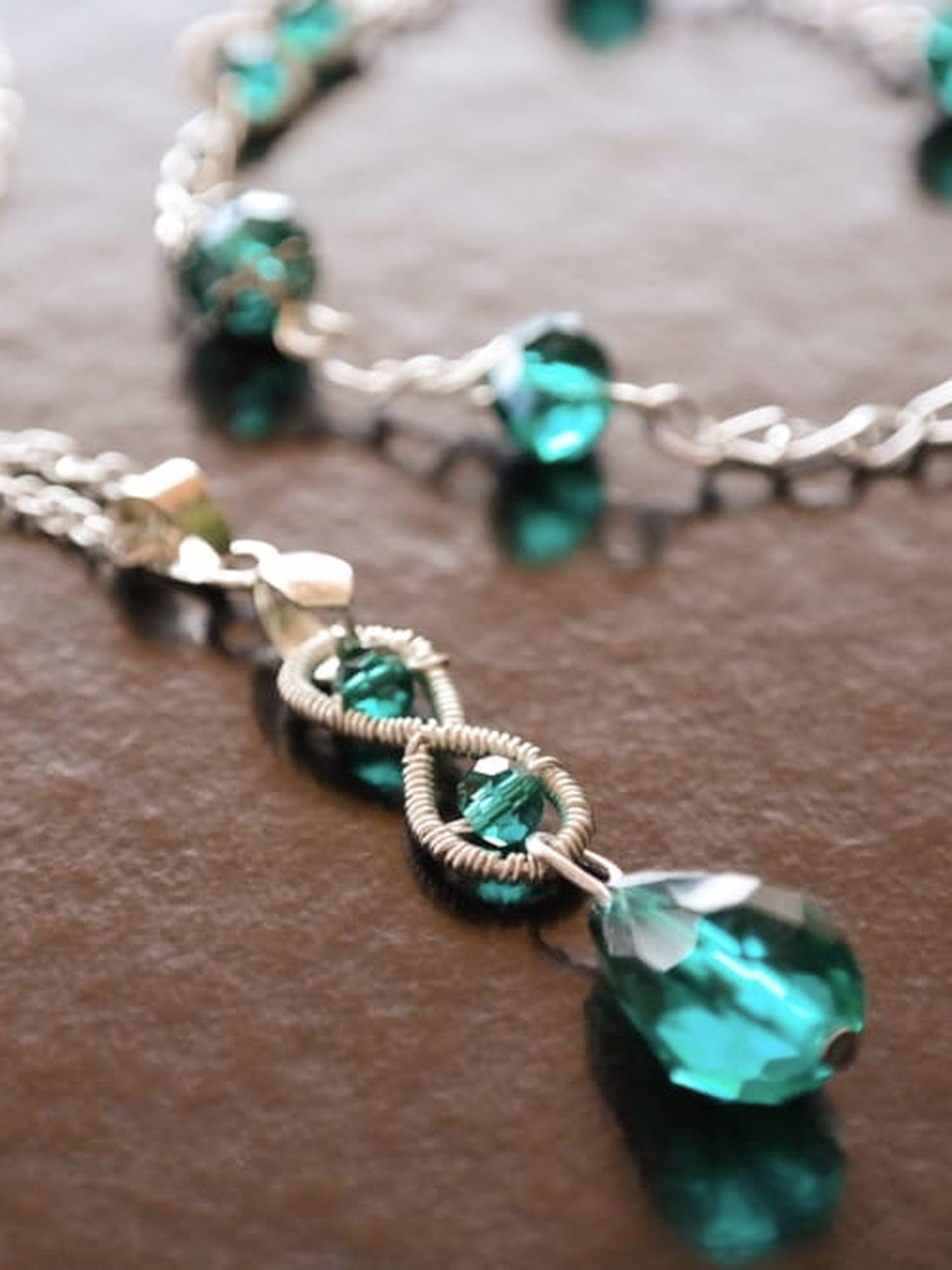 Emerald green teardrop crystal pendant with a matching silver chain bracelet and faceted beads on a dark brown background