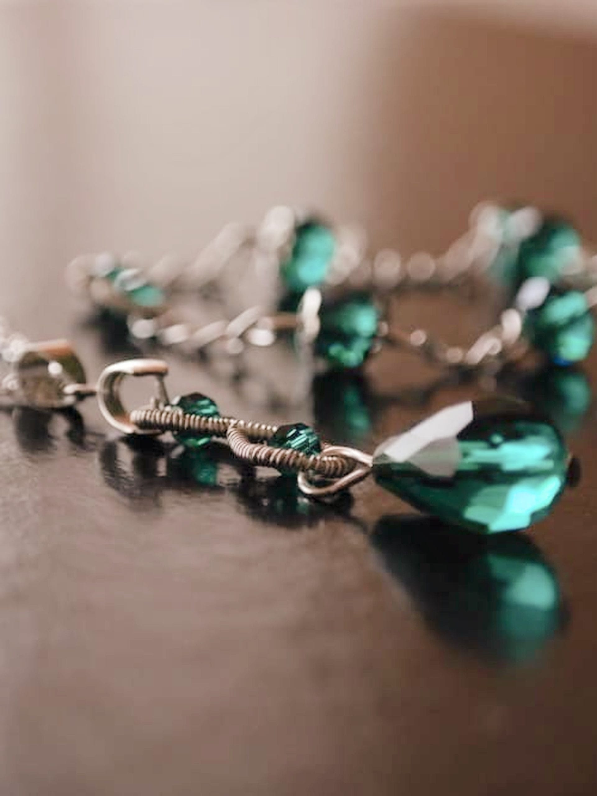 Close-up of a silver wire-wrapped pendant with an emerald green teardrop crystal on a glossy surface, with the matching bracelet blurred in the background