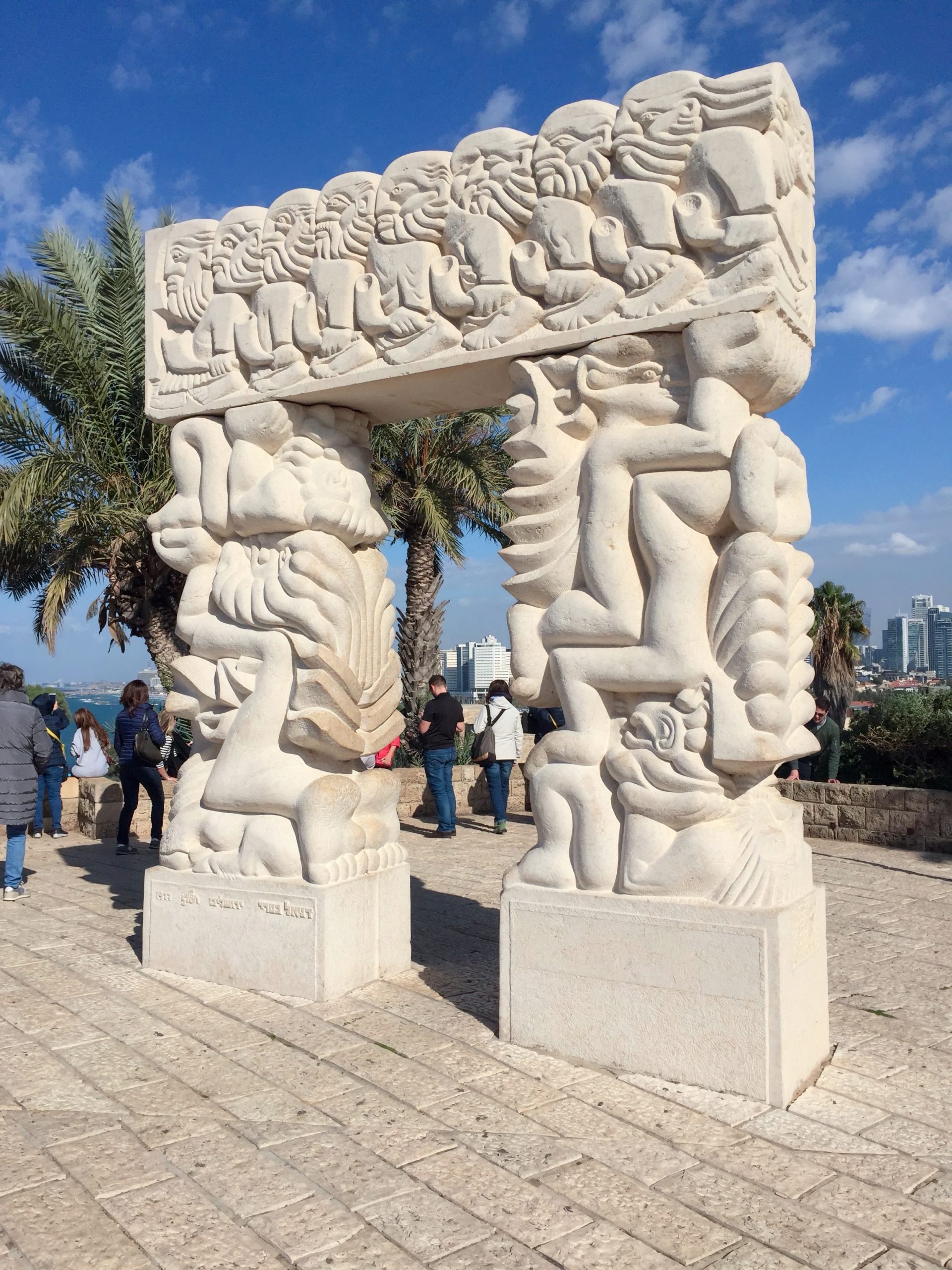 Poarta Credinței din Jaffa, sculptură din piatră deschisă la culoare, cu figuri &icirc;n relief pe două coloane și o grindă, &icirc;ncadrată de palmieri