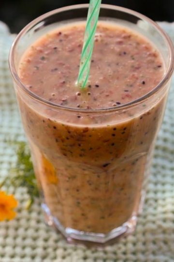 Pahar cu smoothie și pai verde, pe un material verde deschis, lângă o floare galbenă