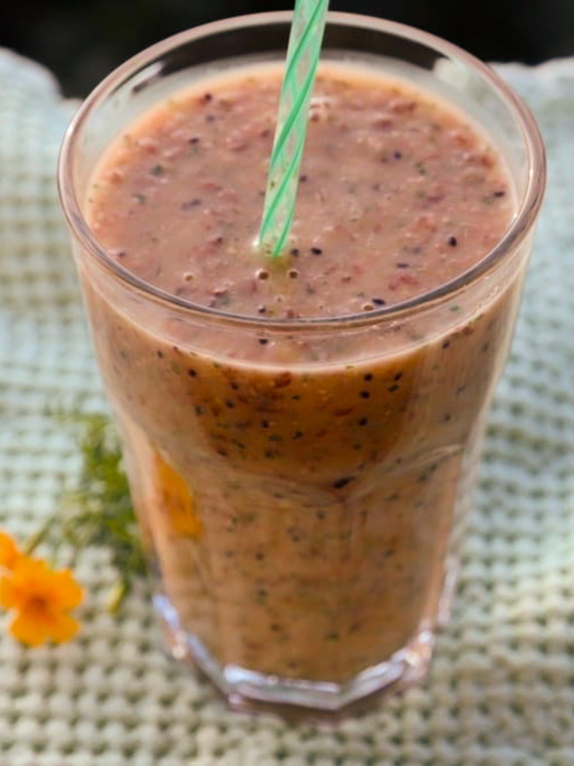 Pahar cu smoothie și pai verde, pe un material verde deschis, lângă o floare galbenă