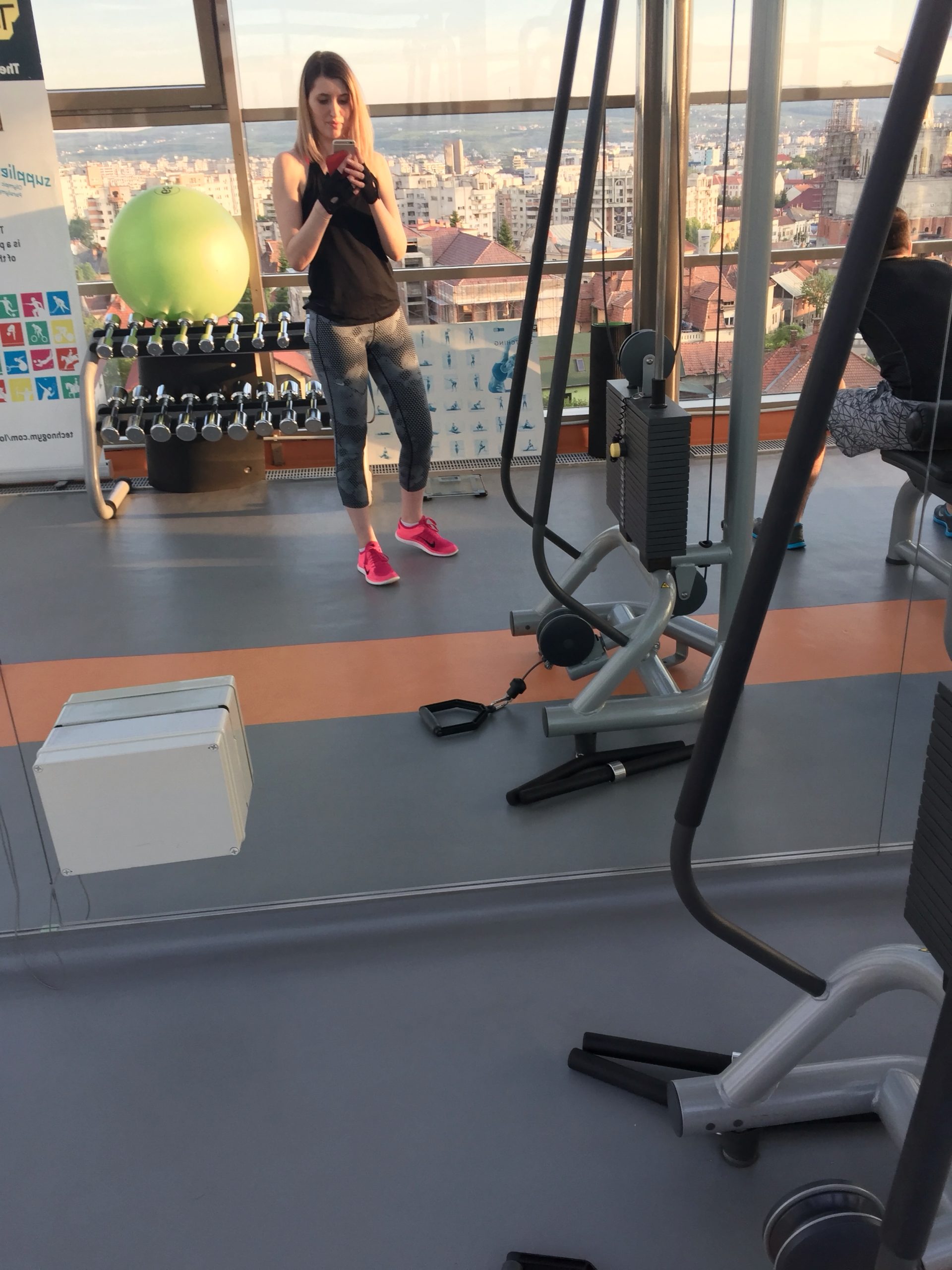 Selfie în oglindă la sala de fitness, lângă suportul cu gantere, cu orașul vizibil prin ferestrele mari