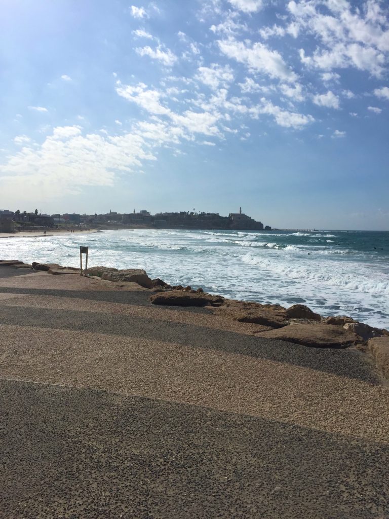 Faleză mărginită de st&acirc;nci, cu valurile Mediteranei și clădirile vechi din Jaffa &icirc;n depărtare