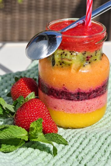 A jar of colourful layered smoothie with a spoon and straw, beside strawberries and mint leaves