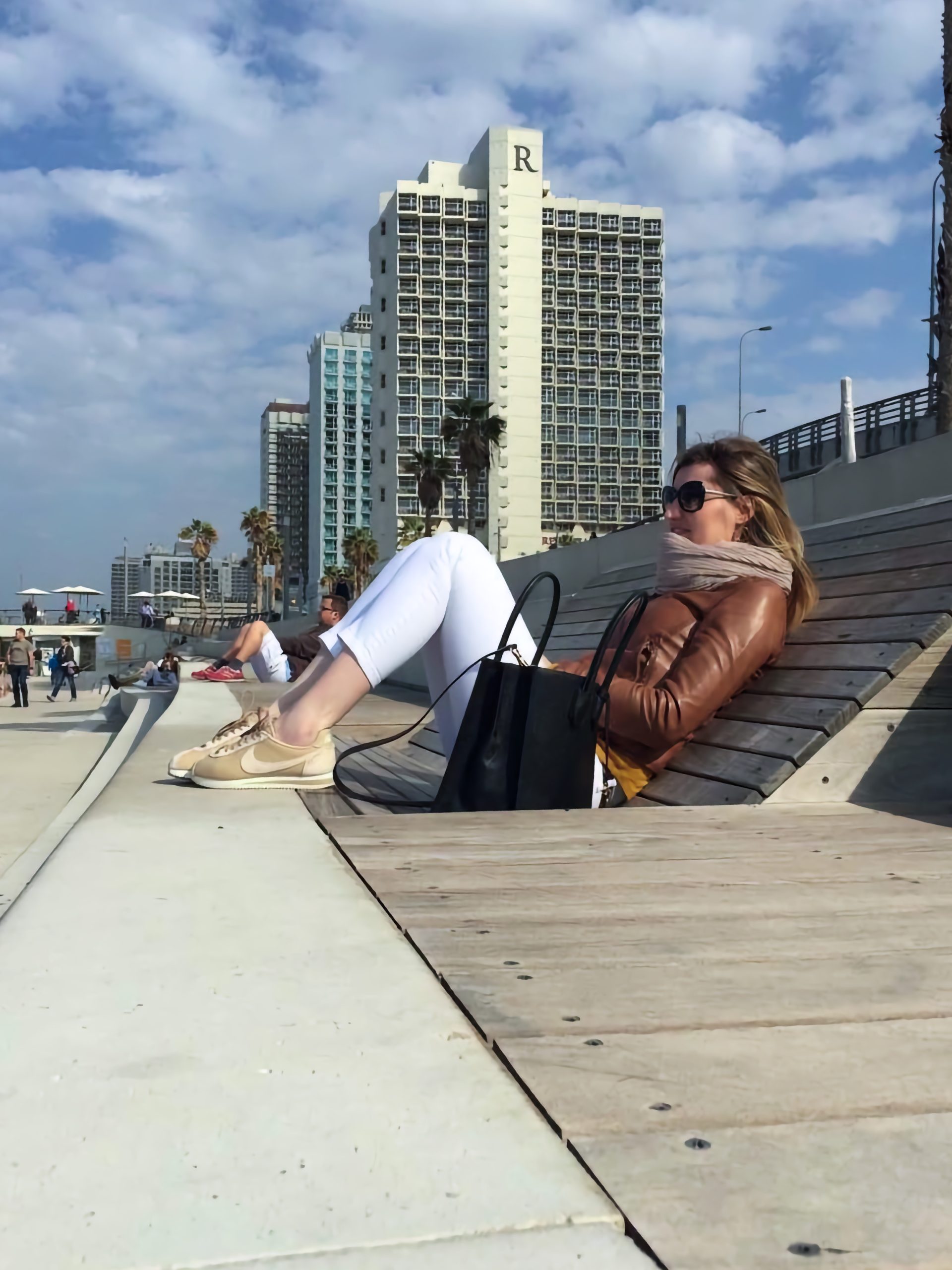 Loredana așezată pe o bancă din lemn pe faleza din Tel Aviv, cu palmieri și clădiri &icirc;nalte &icirc;n fundal