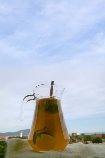 Glass pitcher of lemonade with citrus slices and mint on a green cloth, with sky and distant hills in the background