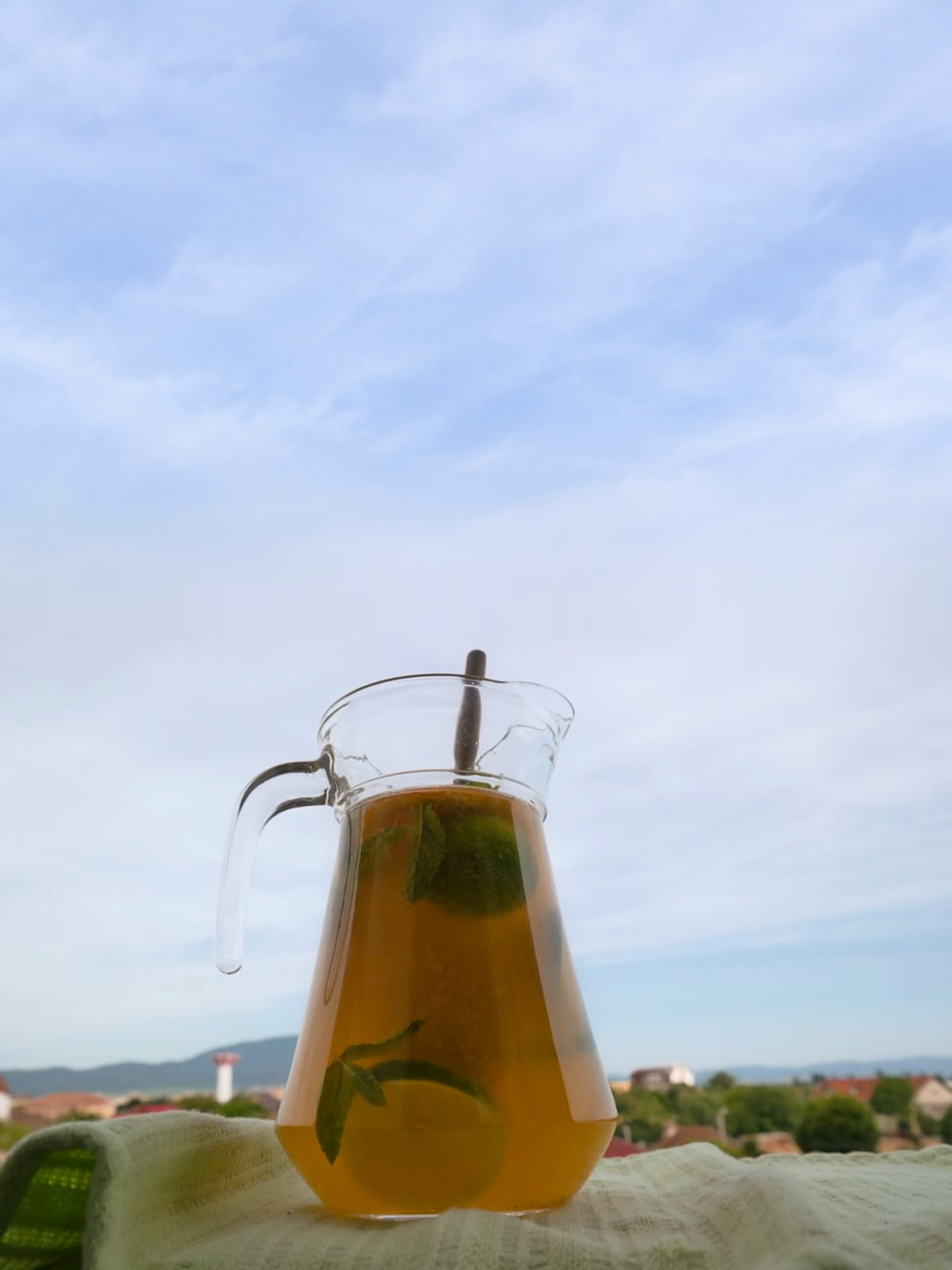 Carafă de sticlă cu limonadă, felii de citrice și mentă, pe un material verde, cu cerul și dealurile &icirc;n fundal