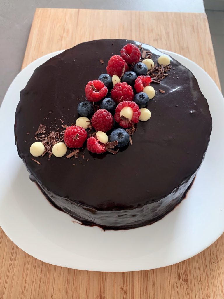 Mixed berry mousse cake with glossy cocoa mirror glaze, decorated with raspberries, blueberries, white chocolate pearls and dark chocolate shavings