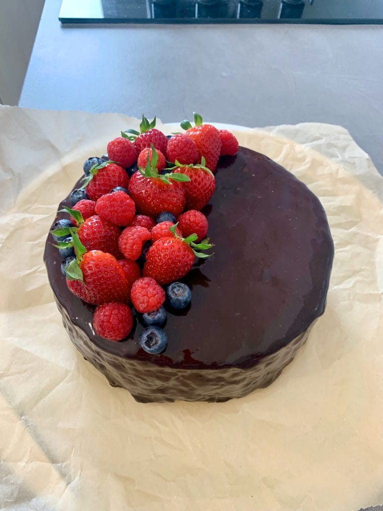 Mixed berry mousse cake covered in glossy cocoa mirror glaze and topped with fresh strawberries, raspberries and blueberries