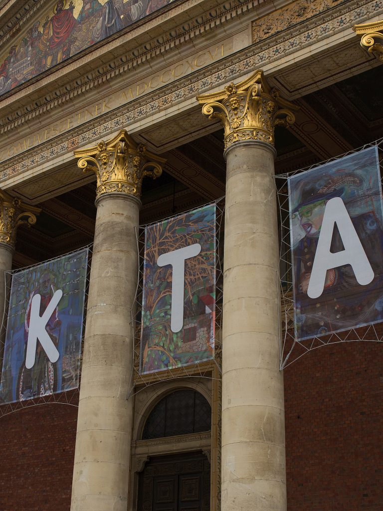 Columns with gilded capitals beneath a mosaic at the banner-covered entrance to Műcsarnok