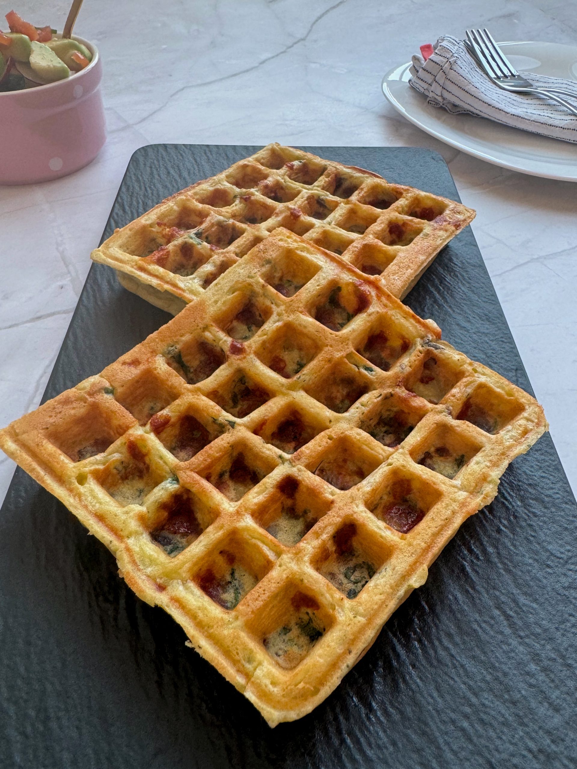 Două gofre rumenite cu ciuperci, mozzarella și spanac, pe un platou negru, cu un bol de salată în fundal