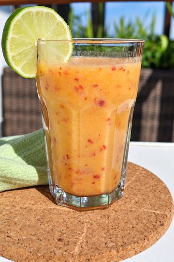 Pahar cu smoothie de piersici și o felie de lime pe margine, pe un suport de plută