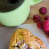 Raspberry honey scone on baking paper beside fresh raspberries and a green cup of coffee