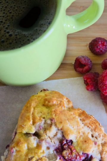 Raspberry honey scone on baking paper beside fresh raspberries and a green cup of coffee