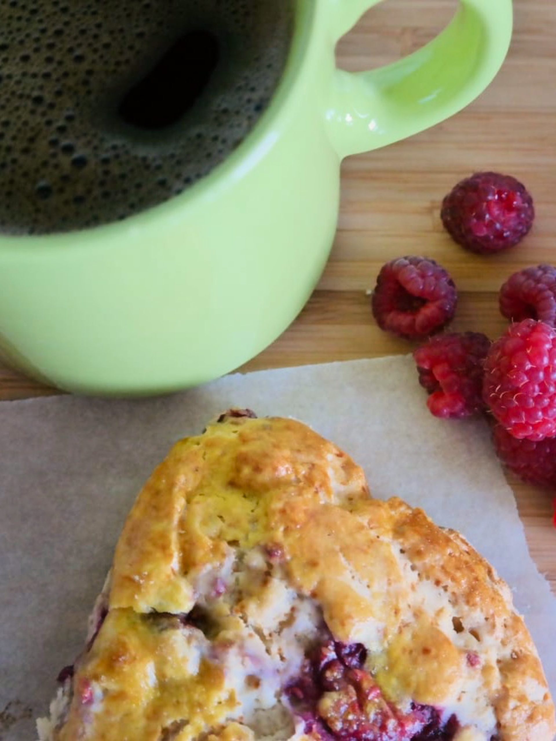 Raspberry honey scone on baking paper beside fresh raspberries and a green cup of coffee