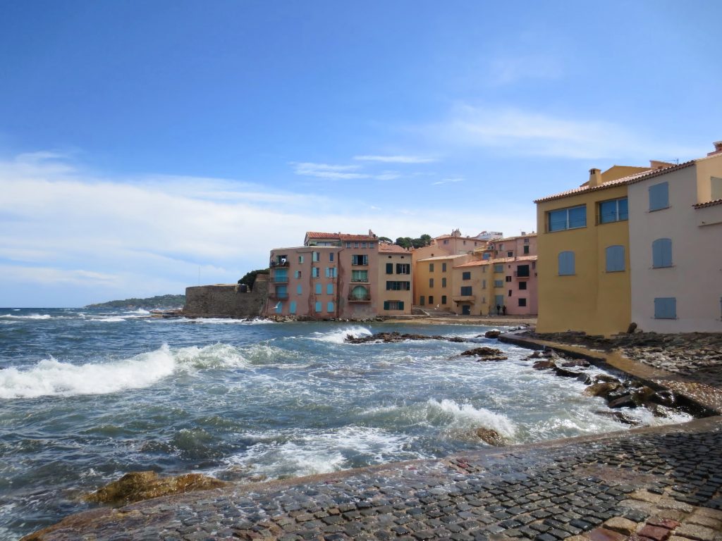 Valuri care se sparg de st&acirc;nci &icirc;n fața caselor colorate de pe malul mării din Saint-Tropez
