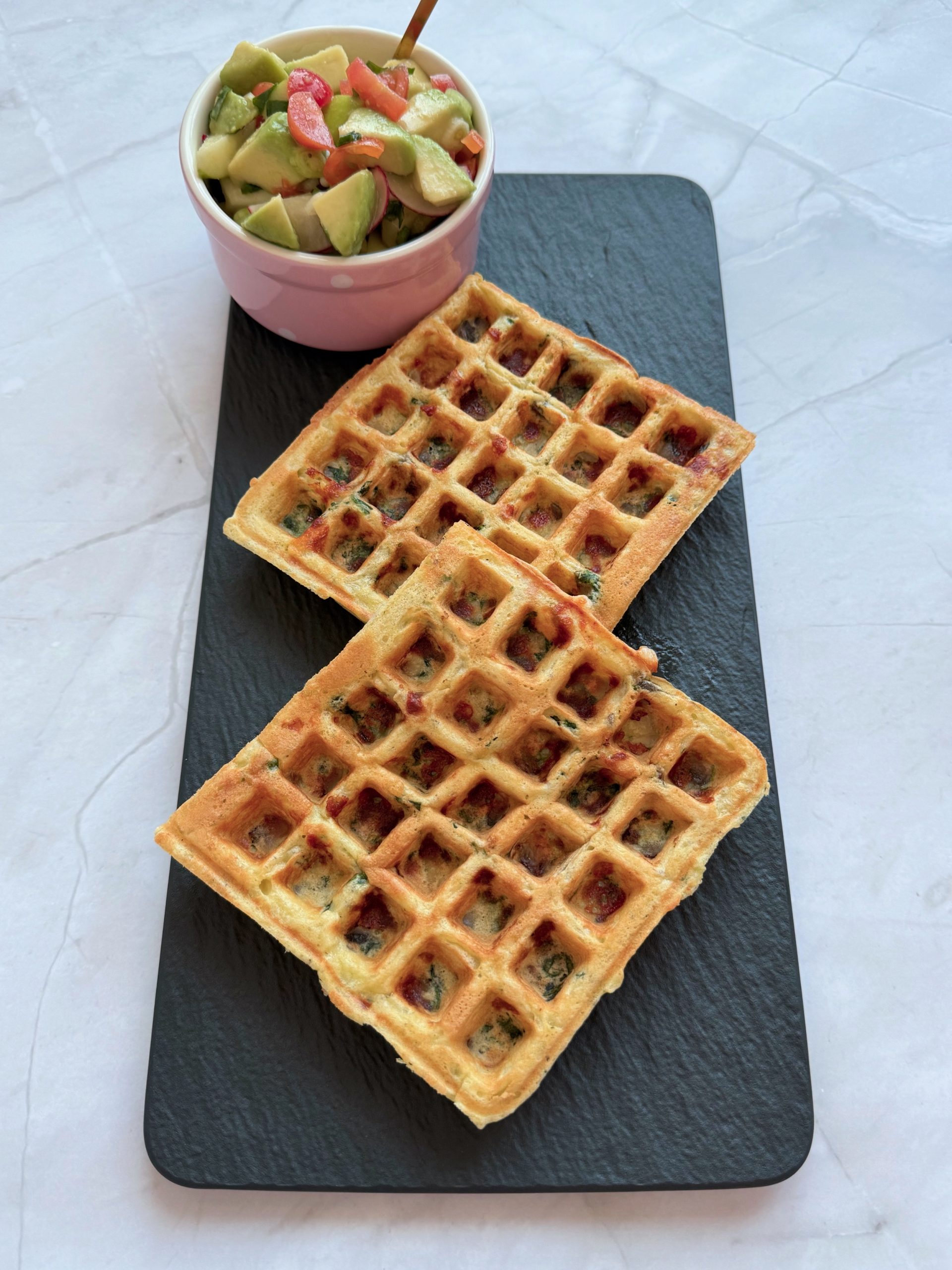 Două gofre cu ciuperci, mozzarella și spanac pe un platou negru, alături de un bol roz cu salată de avocado, roșii, ceapă verde și ridichi