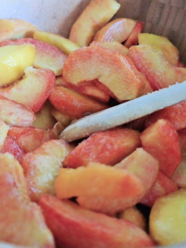 Sliced peaches being mixed with a wooden spoon in a wooden bowl for the cobbler filling