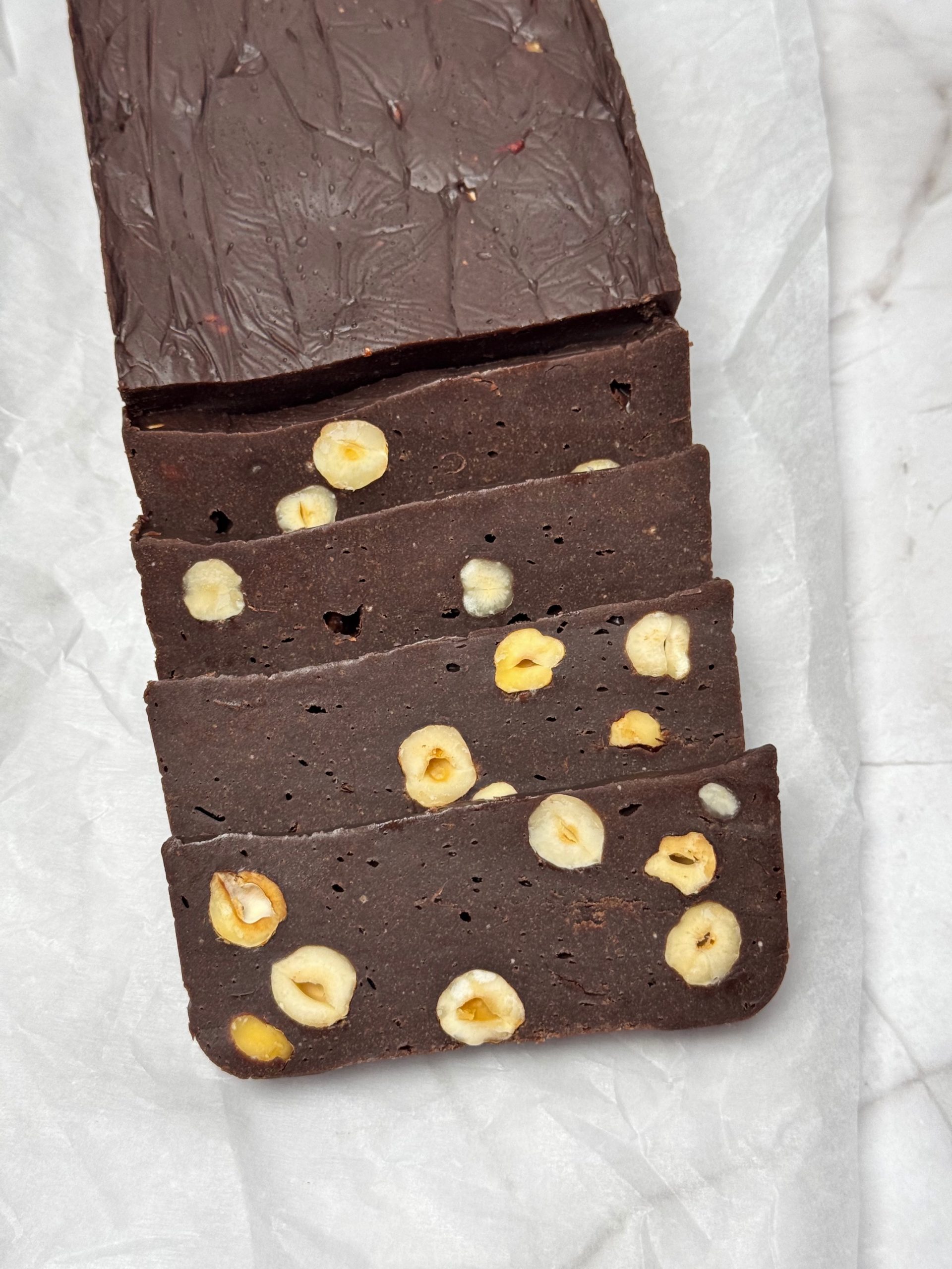 Partially sliced homemade chocolate with four hazelnut slices arranged on white paper