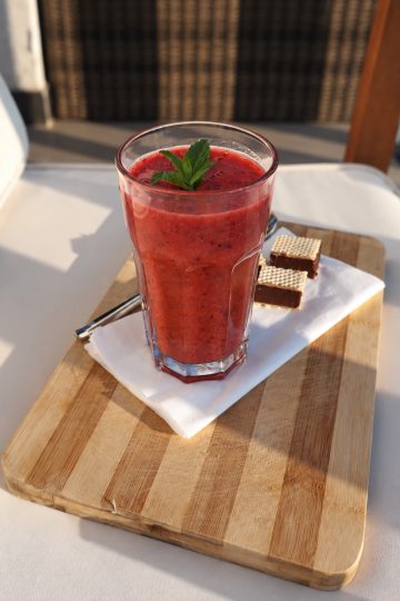 Pahar cu smoothie roșu și frunze de mentă, pe un tocător de lemn, lângă două bucăți de napolitană