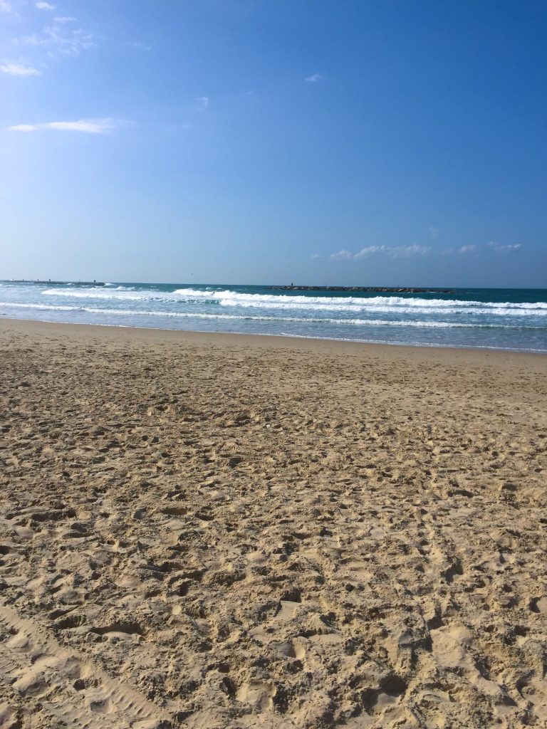 Plajă cu nisip și urme de pași &icirc;n Tel Aviv, cu valuri albe și diguri &icirc;n larg, sub cer albastru