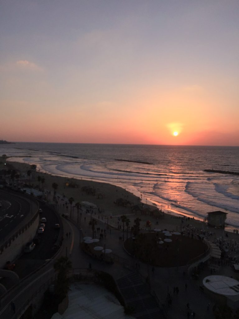Soare deasupra orizontului și reflexii portocalii pe valuri, cu plaja și faleza din Tel Aviv &icirc;n prim-plan