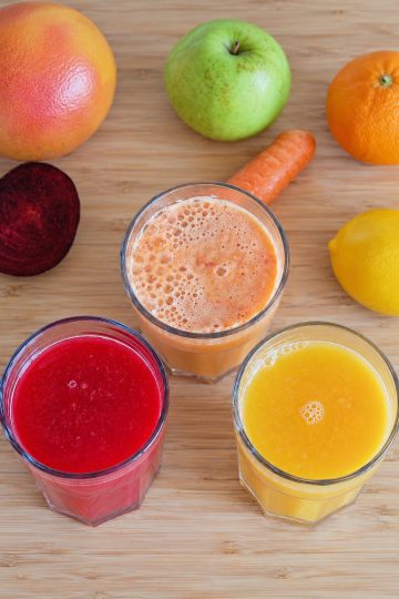 Three glasses of red, pale orange and yellow juice on a wooden surface with fruit and vegetables