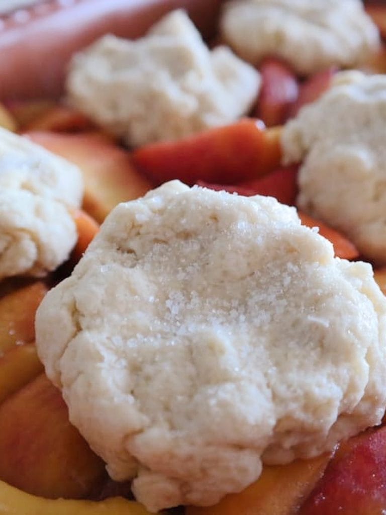 Dollops of raw biscuit dough sprinkled with sugar on top of sliced peaches in a terracotta dish before baking