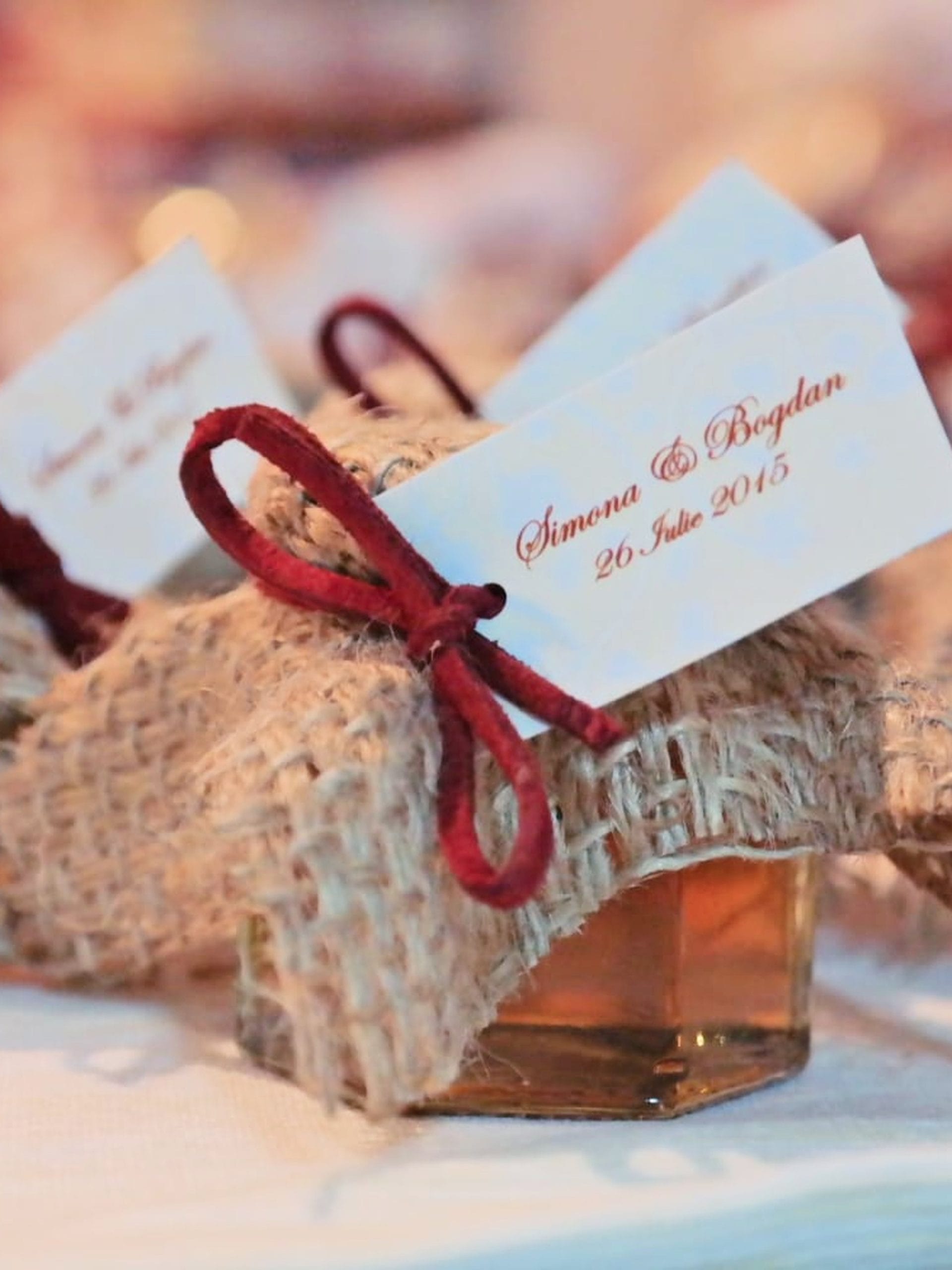 Several small honey jars decorated with burlap, burgundy cord and personalized tags, arranged on a table as wedding favors