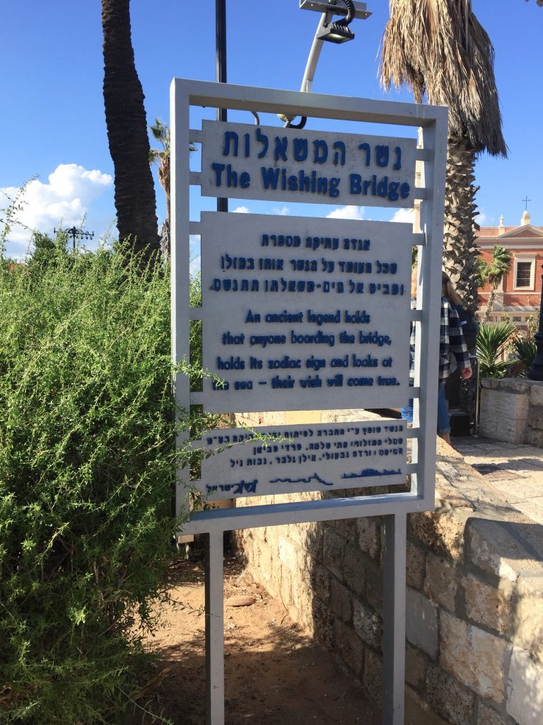 Panou &icirc;n ebraică și engleză cu numele și legenda Podului Dorințelor din Jaffa, l&acirc;ngă palmieri și trepte din piatră